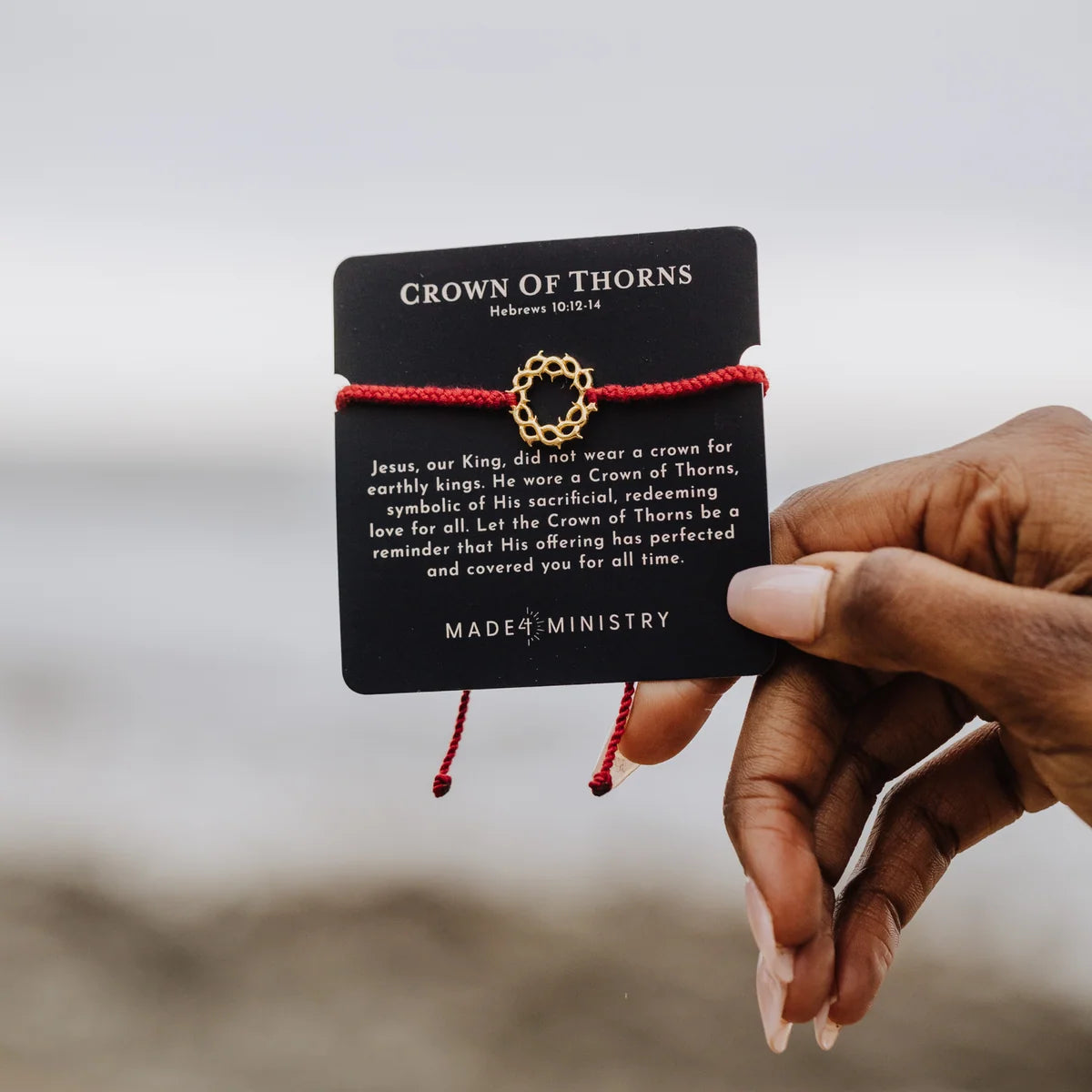 A hand holds a black card displaying a red cord bracelet with a gold crown of thorns charm. The card reads 'CROWN OF THORNS' and 'MADE BY MINISTRY'.