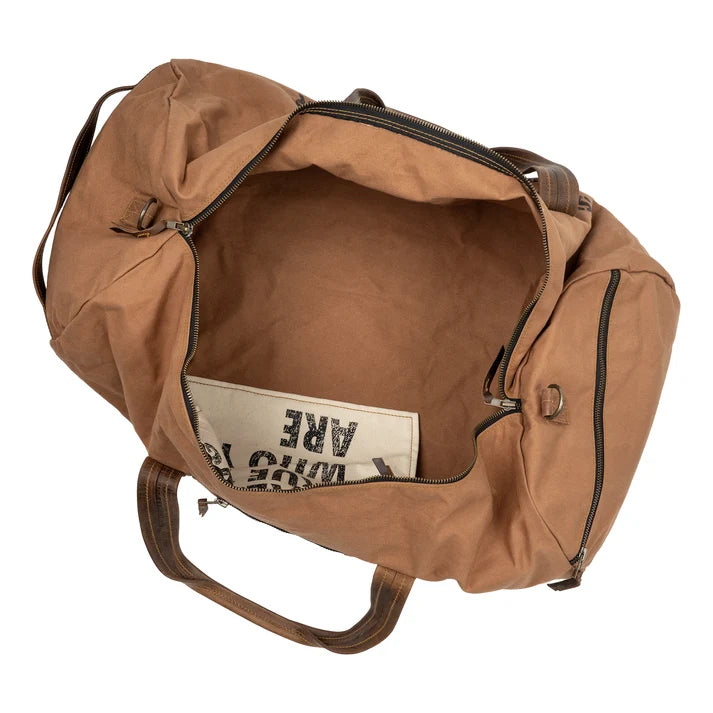 A tan canvas duffel bag shown from an overhead angle, with the main compartment open to reveal a cream-colored interior pocket containing a book or magazine.