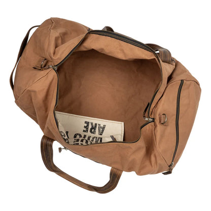 A tan canvas duffel bag shown from an overhead angle, with the main compartment open to reveal a cream-colored interior pocket containing a book or magazine.
