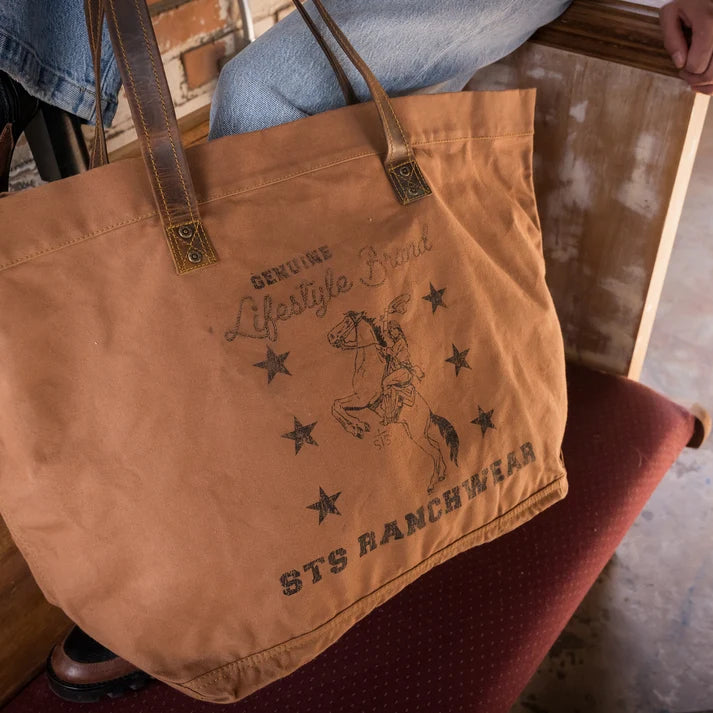 A tan canvas tote bag with brown leather handles and dark printed text reading 'DENUSE Alstyle Brand' and 'STS RANCH WEAR,' shown being carried indoors near a brick wall and chair.