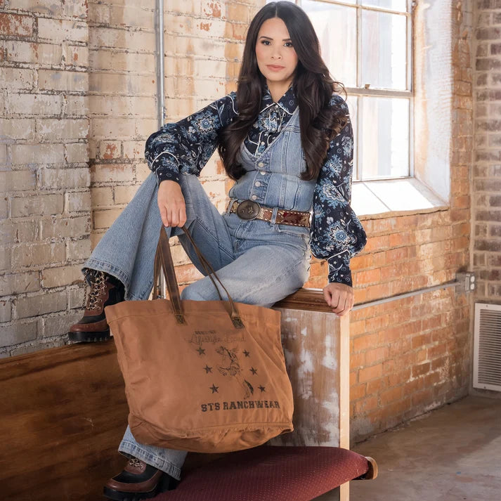 A woman sits on a wooden bench in a rustic brick room, holding a large brown canvas tote bag with 'STS RANCHHOUSE' printed on it. She is wearing a blue floral long-sleeve shirt, denim overalls, and brown boots.