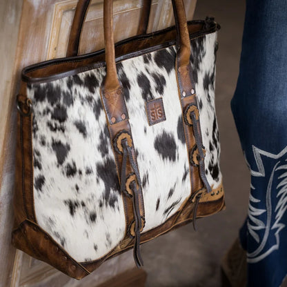 A cowhide tote bag with brown leather trim and handles, featuring a small square SIS logo patch on the front, shown hanging against a wooden door next to a person's jeans.