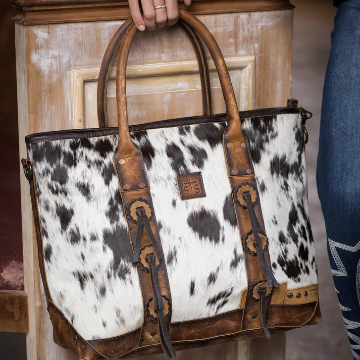 A cowhide tote bag with brown leather handles and trim, held by a person in a rustic indoor setting.