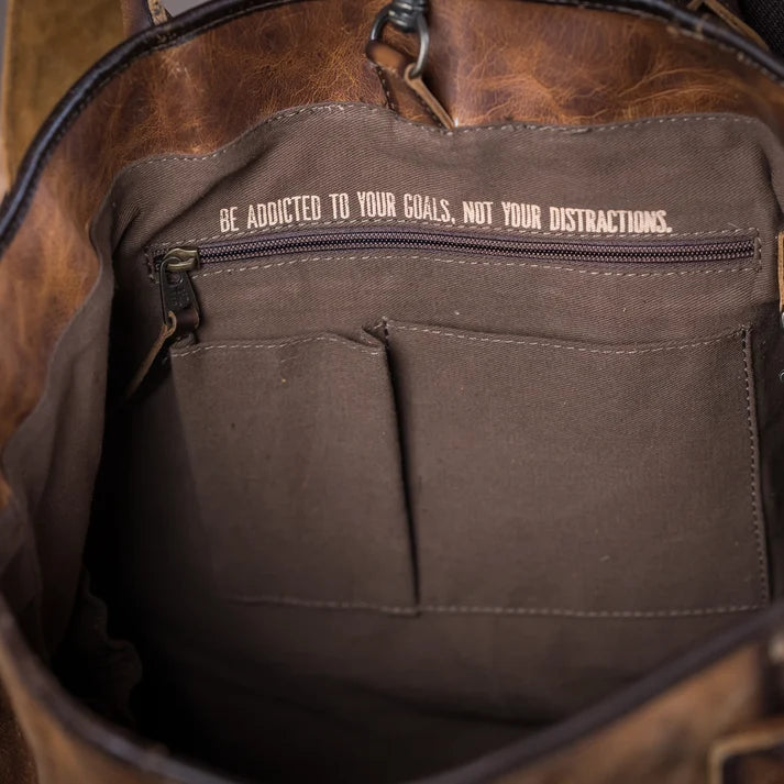 Close-up of the interior of a brown leather bag, showing a dark brown canvas pocket with a zipper and the printed text 'BE ADDICTED TO YOUR GOALS, NOT YOUR DISTRACTIONS.'.