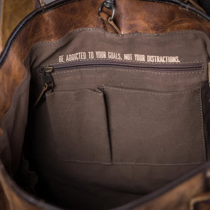 Close-up of the interior of a brown leather bag, showing a dark brown canvas pocket with a zipper and the printed text 'BE ADDICTED TO YOUR GOALS, NOT YOUR DISTRACTIONS.'.