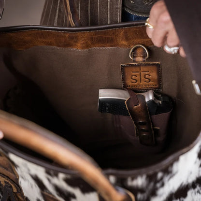 Close-up of the inside of a large brown leather bag with a side pocket holding a passport and small items. A leather patch with the embossed initials 'STS' is visible, and part of a person's hand with rings is seen holding the bag open.