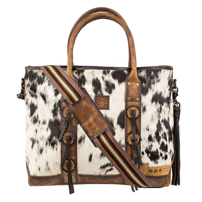 A western-style tote bag featuring a black and white cowhide body, brown leather handles and trim, and a striped leather shoulder strap with tassel details, shown on a plain white background.