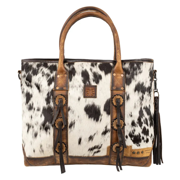 A western-style tote bag featuring a black and white cowhide pattern body with brown leather handles, trim, and decorative strap details, shown on a plain white background.