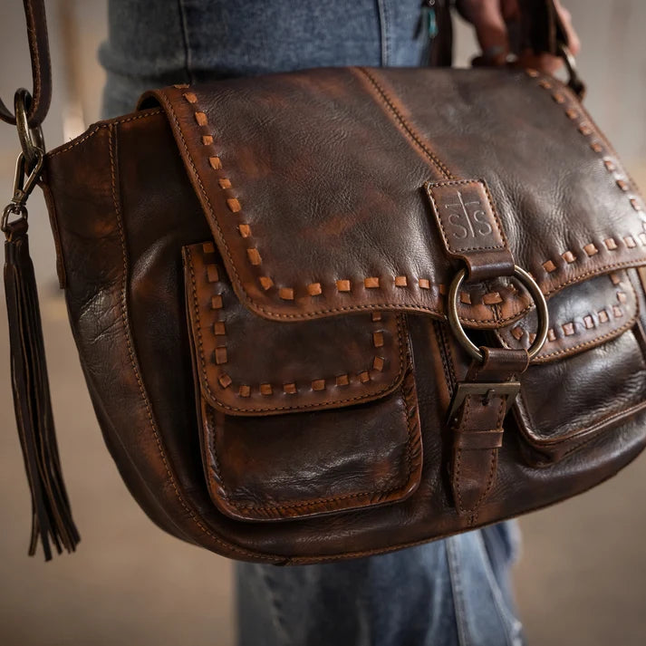 A close-up of a dark brown leather shoulder bag with decorative stitching, a tassel, and a metal ring clasp, being worn by a person in blue jeans.