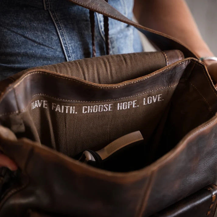 Close-up of a brown leather shoulder bag being held open by a person wearing blue jeans. The inside of the bag is lined with brown fabric printed with the words 'HAVE FAITH.