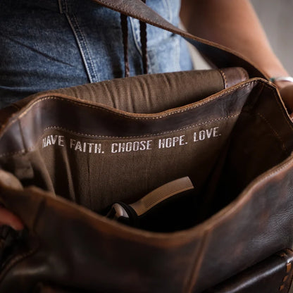 Close-up of a brown leather shoulder bag being held open by a person wearing blue jeans. The inside of the bag is lined with brown fabric printed with the words 'HAVE FAITH.