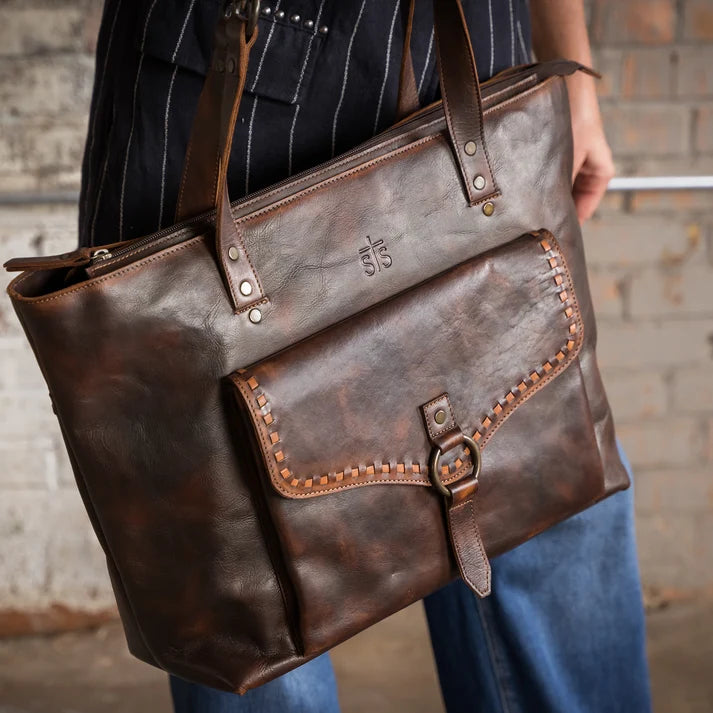 A person holds a large distressed brown leather tote bag with a front pocket and buckle detail, shown against a rustic brick wall background.