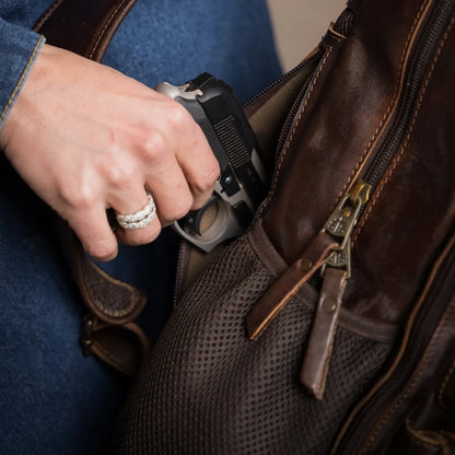 A close-up lifestyle shot of a person's hand reaching into a brown leather and mesh crossbody bag to retrieve a handgun. The person is wearing a blue denim jacket and a silver ring.