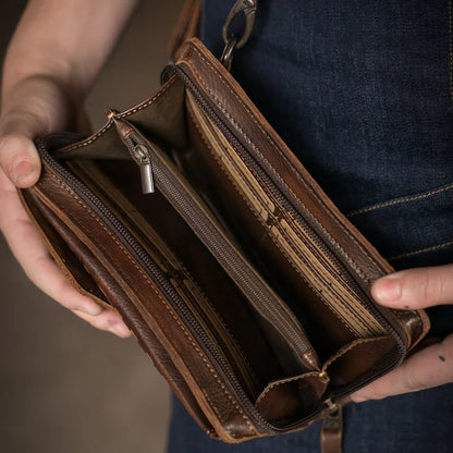 A close-up of a person holding an open brown leather wallet or clutch with multiple zip compartments and card slots, showing the interior construction against a dark denim-clad torso.