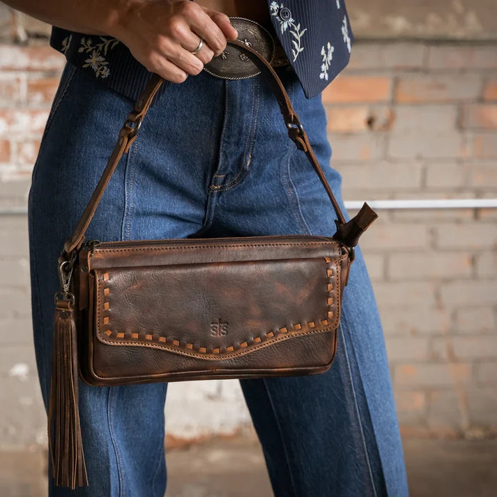 A brown distressed leather crossbody bag with a tassel and decorative studs, worn by a person in blue jeans against a brick wall background.