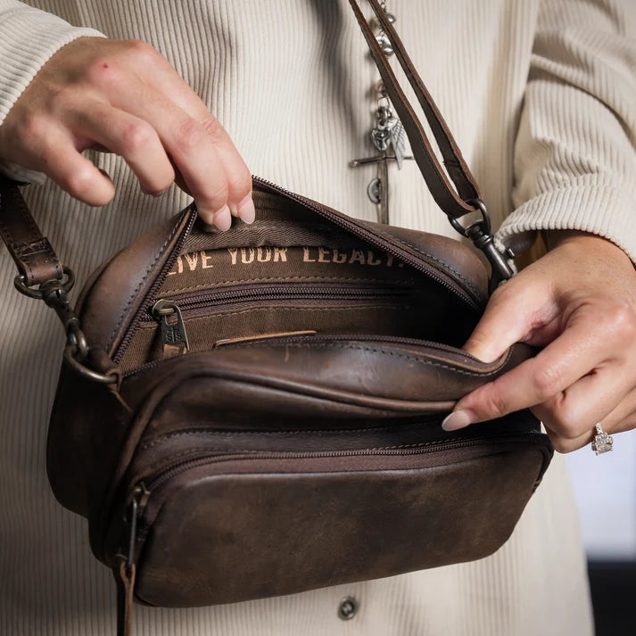 A close-up of a person holding open a dark brown leather crossbody bag, showing the interior compartments and lining.