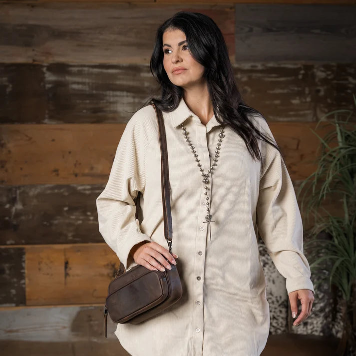 A woman wearing a cream-colored long-sleeve button-up shirt and a long beaded necklace, holding a dark brown leather crossbody bag, set against a rustic wooden wall background.