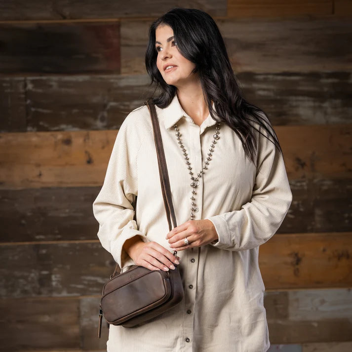 A woman with long dark hair wearing a cream-colored button-up shirt and a long beaded necklace, carrying a brown leather crossbody bag against a rustic wooden plank wall.