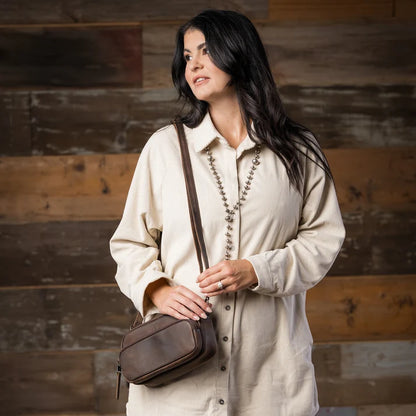 A woman with long dark hair wearing a cream-colored button-up shirt and a long beaded necklace, carrying a brown leather crossbody bag against a rustic wooden plank wall.