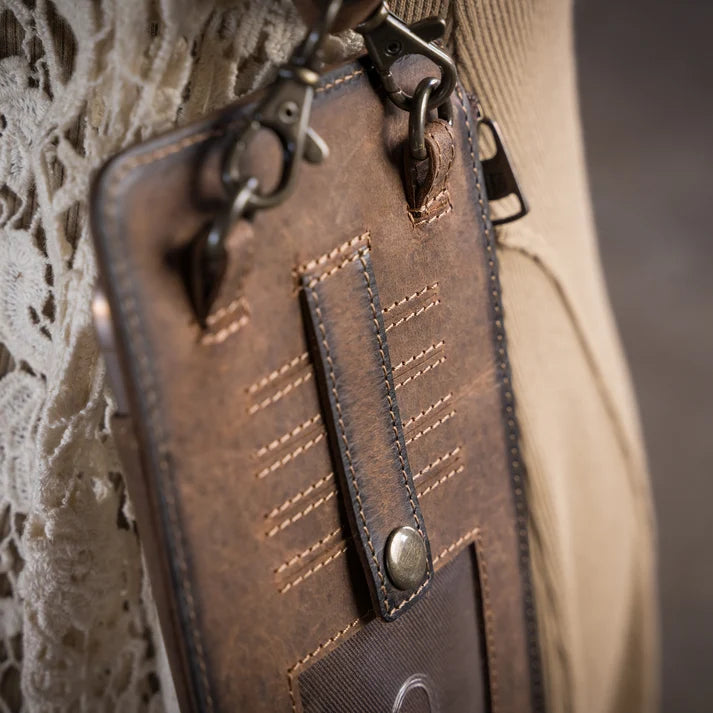 A close-up of a brown distressed leather phone pouch or card holder with metal clips and a snap-button strap, shown hanging against a beige knit garment and lace fabric background.