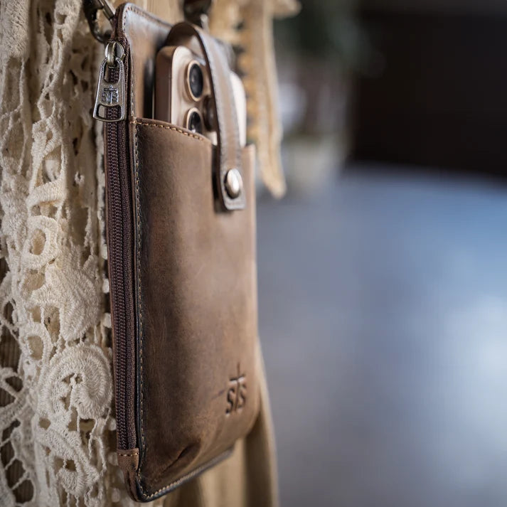 A close-up of a brown leather phone pouch with a zipper and snap closure, holding a smartphone, hanging against a cream lace garment.