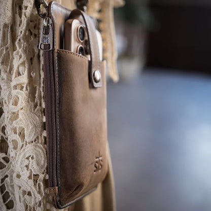 A close-up of a brown leather phone pouch with a zipper and snap closure, holding a smartphone, hanging against a cream lace garment.