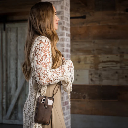 A woman in a beige dress wearing a cream lace cardigan and carrying a brown leather crossbody phone pouch, standing in front of a rustic wooden and brick wall.