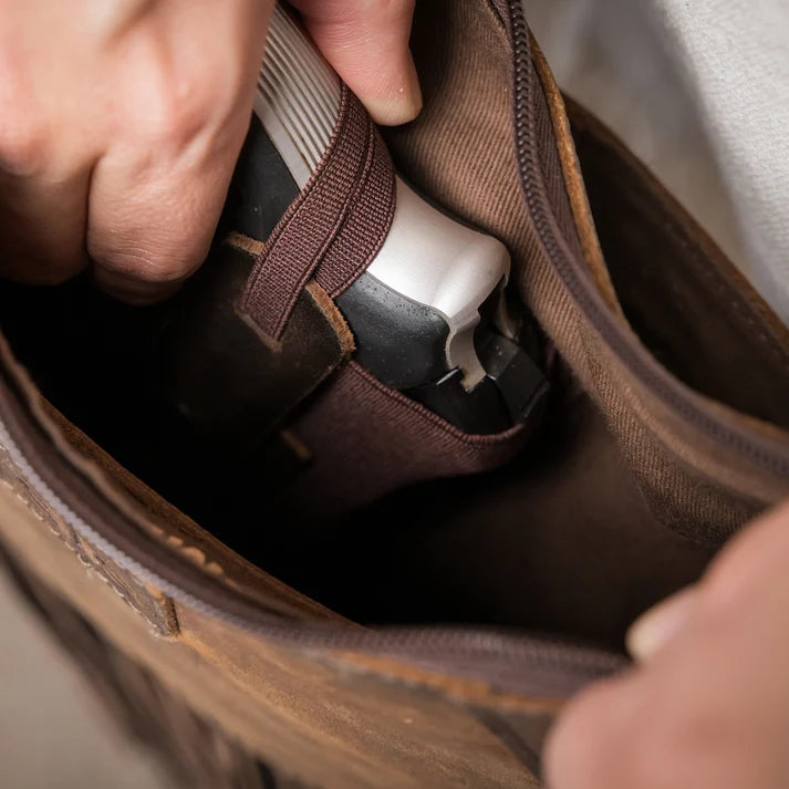 A close-up of a person's hands placing a brown leather and canvas shoe bag into a brown leather jacket pocket. The bag features a brown elastic strap and a dark brown interior.