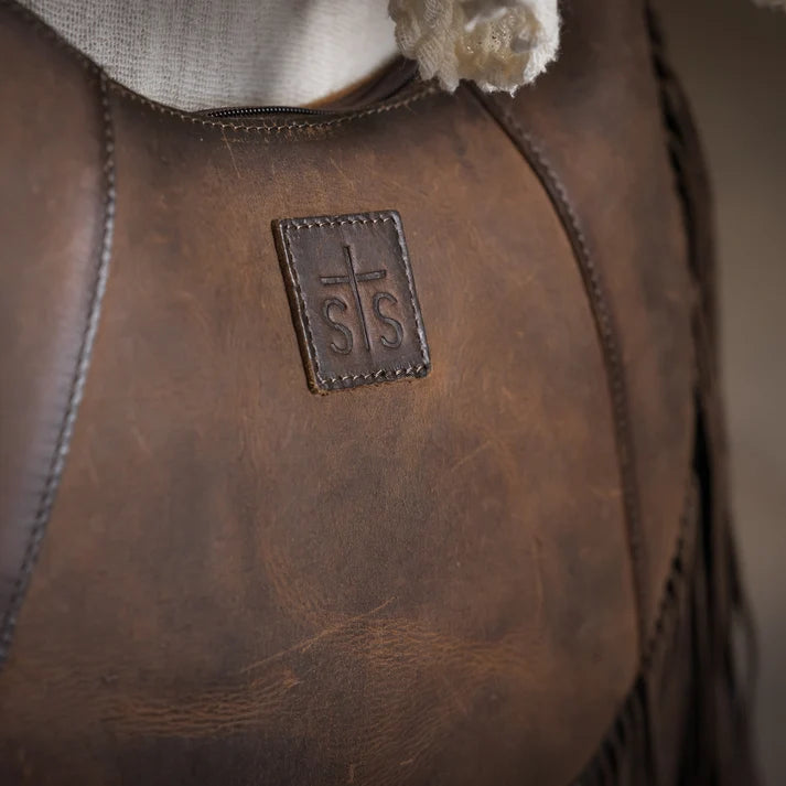 Close-up of a brown leather shoulder bag with fringe, showing a stitched square patch with a cross and the letters 'S.S.'
