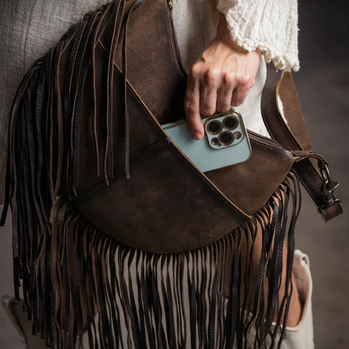 A close-up of a dark brown leather fringe bag being worn crossbody, with a person's hand reaching into the front pocket to insert a light blue smartphone. The bag features long hanging fringe and a wide shoulder strap, styled with a cream-colored textured top and white ankle boots.