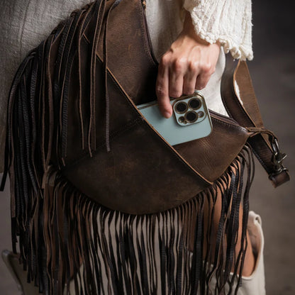 A close-up of a dark brown leather fringe bag being worn crossbody, with a person's hand reaching into the front pocket to insert a light blue smartphone. The bag features long hanging fringe and a wide shoulder strap, styled with a cream-colored textured top and white ankle boots.