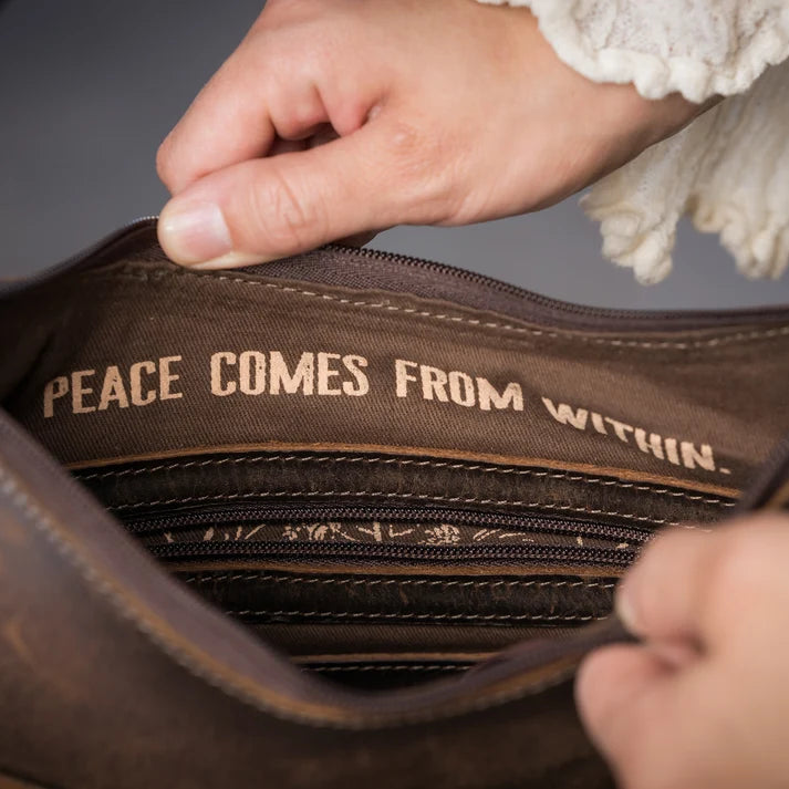 Close-up of a brown leather or faux-leather bag being opened by hands, with the interior lining showing the text 'PEACE COMES FROM WITHIN.' in tan lettering.