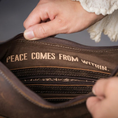 Close-up of a brown leather or faux-leather bag being opened by hands, with the interior lining showing the text 'PEACE COMES FROM WITHIN.' in tan lettering.