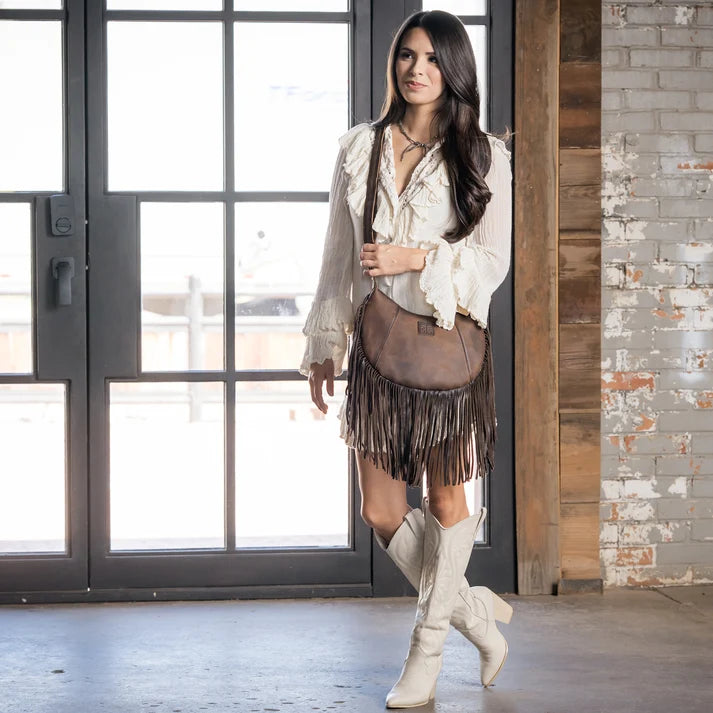 A woman stands indoors wearing a bohemian-style outfit featuring a white ruffled blouse, a brown fringe sash bag, a brown fringe mini skirt, and off-white knee-high boots. She is posed in front of a large window and a brick wall.