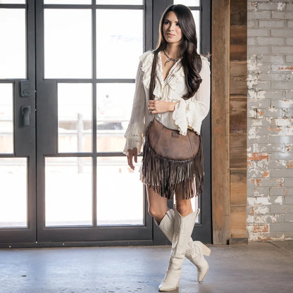 A woman stands indoors wearing a bohemian-style outfit featuring a white ruffled blouse, a brown fringe sash bag, a brown fringe mini skirt, and off-white knee-high boots. She is posed in front of a large window and a brick wall.