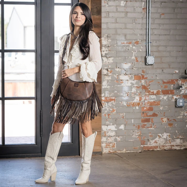 A woman stands in a loft-style room with a brick wall and glass doors, wearing a white fringed bohemian-style top, a brown leather crossbody bag with fringe, and tall white cowboy boots.