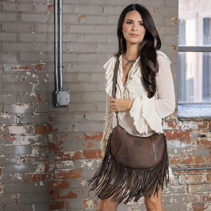 A woman models a brown leather fringe bag with a small gold logo plaque, styled with a cream ruffled blouse and a brown fringe skirt, standing in front of a rustic brick wall.