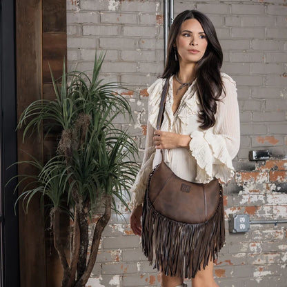 A woman wearing a cream ruffled blouse and a brown leather fringe crossbody bag stands in front of a brick wall next to a potted plant.