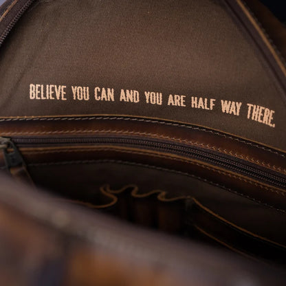 Close-up of the inside of a brown leather bag showing a zipper, stitching, and tan embroidered text that reads, 'BELIEVE YOU CAN AND YOU ARE HALF WAY THERE.'.