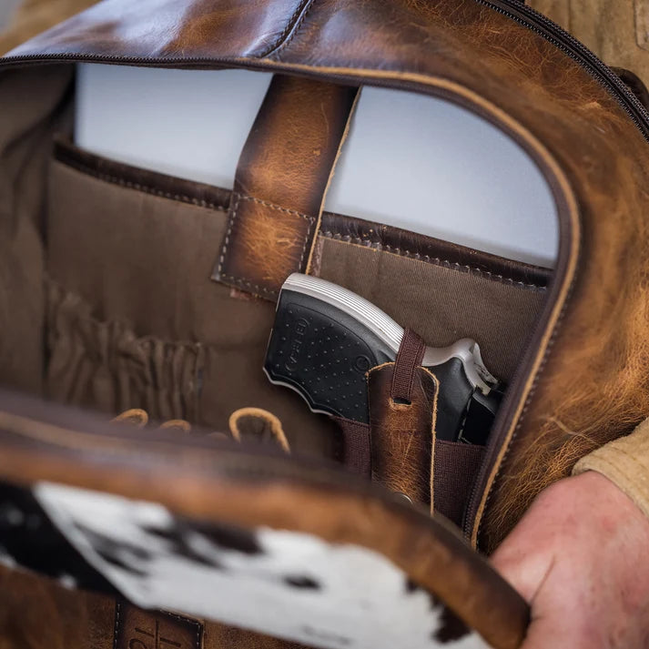 Close-up of an open brown leather bag showing its interior lining, a side pocket, and a handgun tucked inside. A hand is visible holding the bag open.