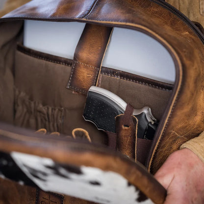 Close-up of an open brown leather bag showing its interior lining, a side pocket, and a handgun tucked inside. A hand is visible holding the bag open.