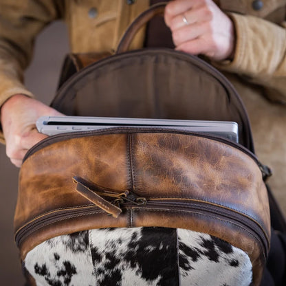 A close-up of a brown leather backpack with a cowhide lower panel, shown being held open by a person wearing a tan jacket. A silver laptop is tucked into the main compartment.