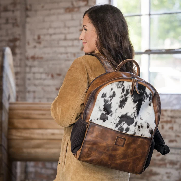 A woman wearing a tan suede jacket is shown from the back, carrying a brown leather and cowhide backpack in a rustic indoor setting with a brick wall and window.