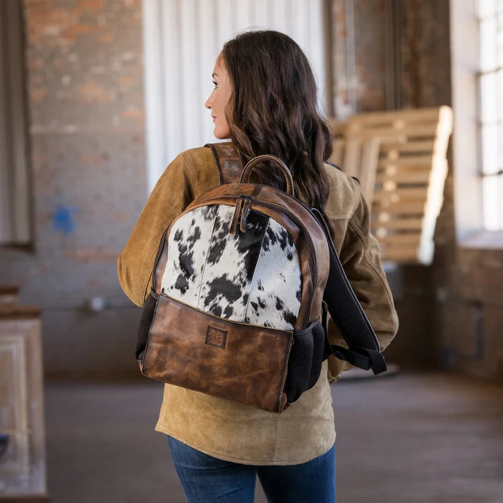 A woman wearing a brown leather and cowhide backpack, photographed from behind in a rustic indoor loft setting.