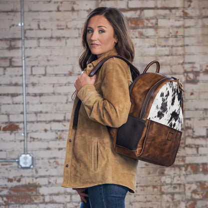 A woman wearing a tan suede jacket and blue jeans carries a brown leather and cowhide backpack against a rustic brick wall background.