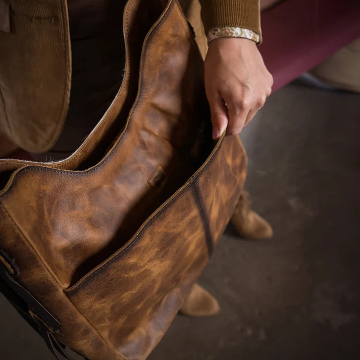 A close-up of a person holding the opening of a large distressed brown leather bag, with part of their brown jacket and hand visible.