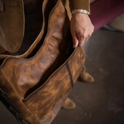 A close-up of a person holding the opening of a large distressed brown leather bag, with part of their brown jacket and hand visible.