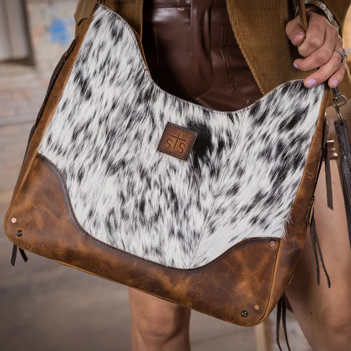 A close-up of a person carrying a large shoulder bag made of cowhide and brown leather. The bag features a black-and-white cowhide upper panel, brown leather trim at the bottom and edges, and a small brown leather logo patch with the initials 's15'.