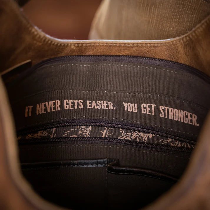 Close-up of the interior of a brown suede or leather bag, showing a dark fabric band with the motivational quote 'IT NEVER GETS EASIER. YOU GET STRONGER.