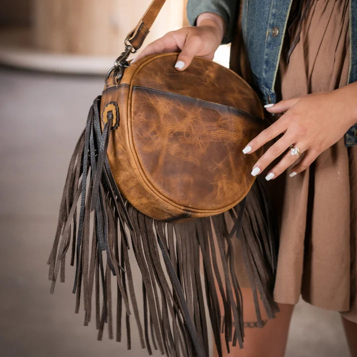 A person holding a round brown distressed leather crossbody bag with long dark brown fringe tassels and a blue denim jacket, wearing a brown dress.